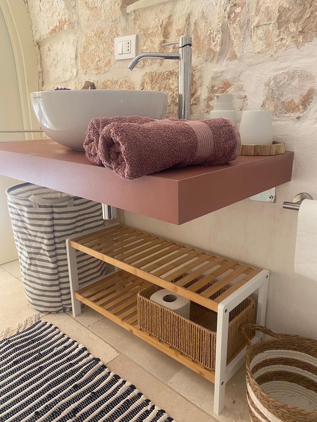 Une salle de bain avec un mur en pierre, un lavabo blanc, une serviette rose et une étagère en bois.