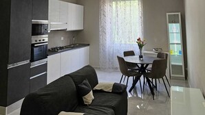 Ett modernt vardagsrum med en svart soffa, ett matbord med stolar, ett kök med vita skåp och ett stort fönster med tunna, genomskinliga gardiner.