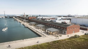 A waterfront building with a covered walkway, a marina with boats, and a large cargo ship in the background. - HENRI Hotel Cuxhaven (Cuxhaven)