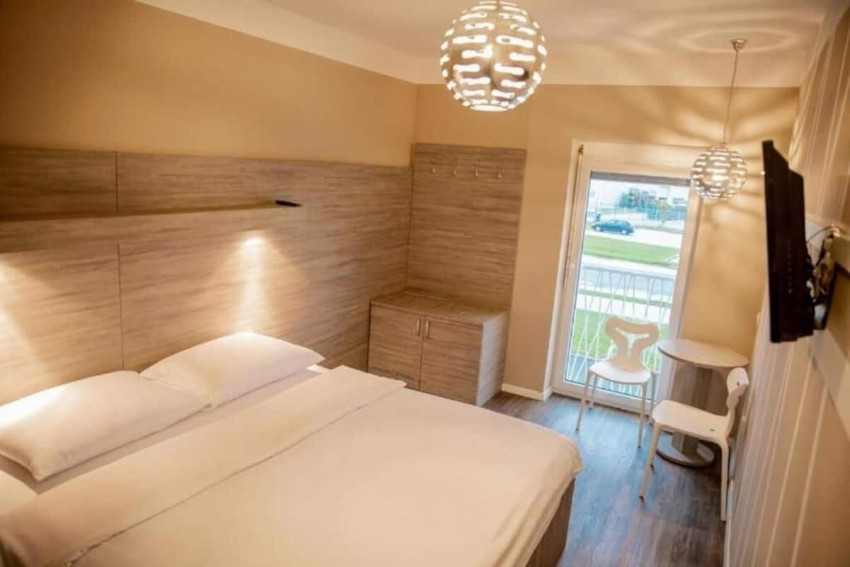 A bedroom with a bed, a small cabinet, and a balcony with a chair.