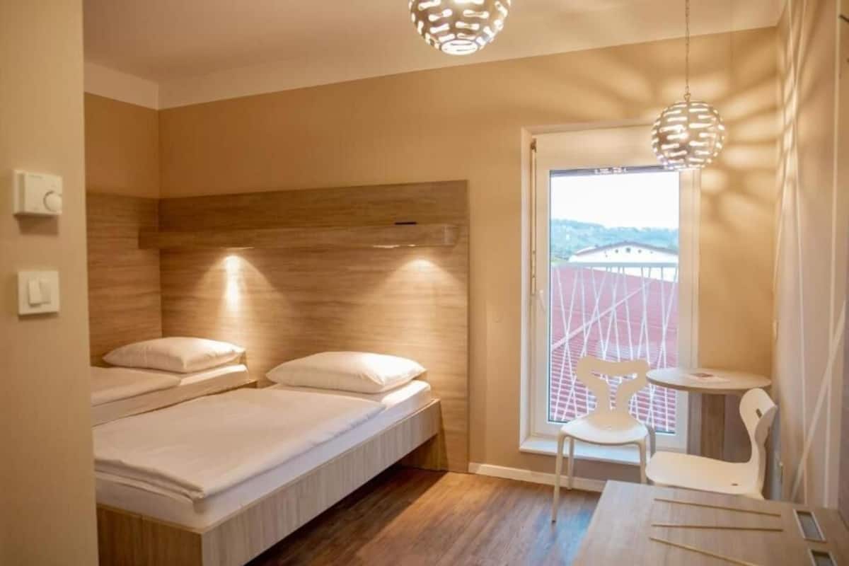 A modern bedroom with two beds, a wooden headboard, a small table, and a chair near a window with a view of buildings.
