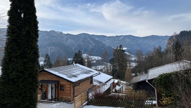 Una cabaña de madera con techo nevado, rodeada de montañas y árboles de hoja perenne.