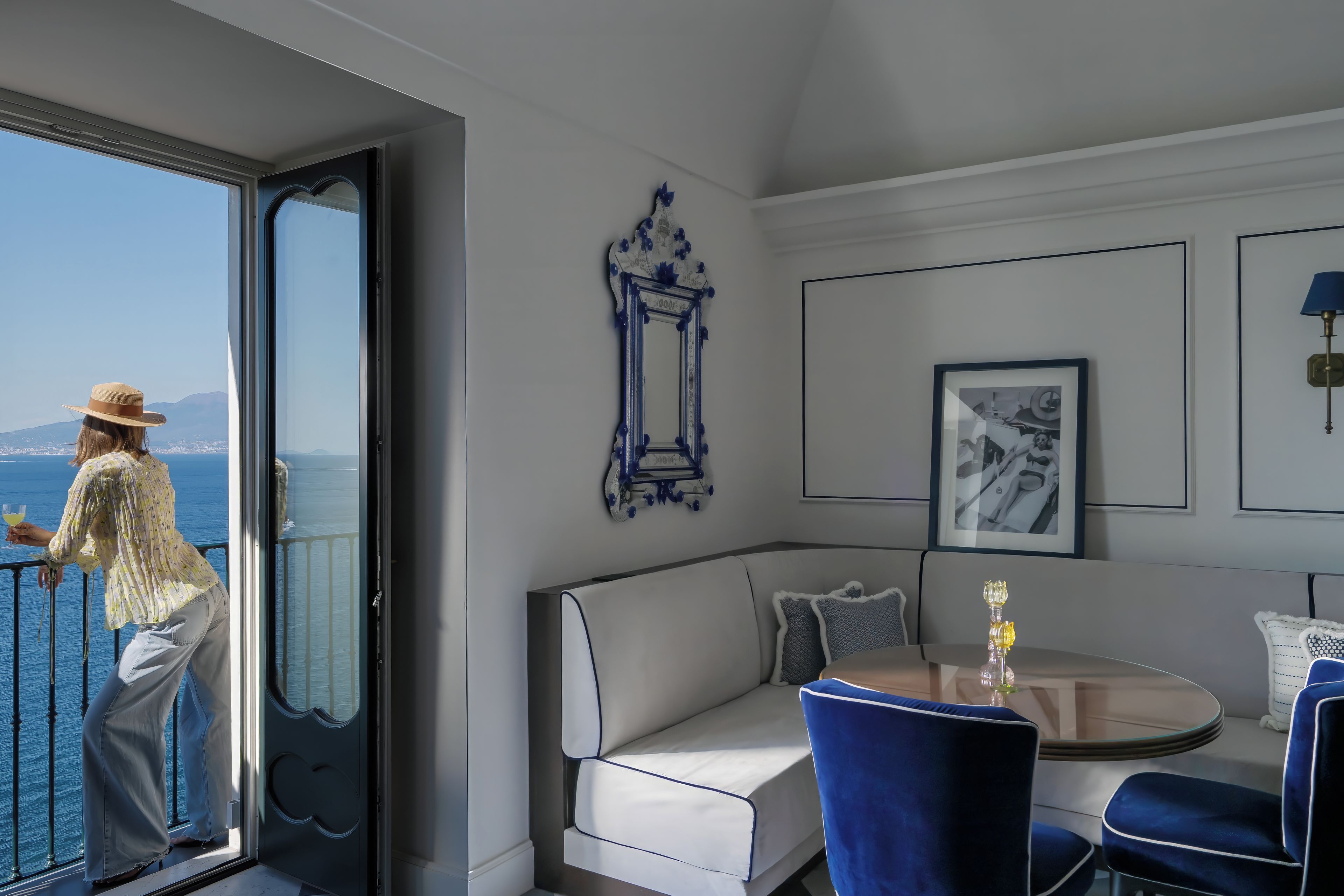 Une chambre avec un balcon donnant sur la mer, un canapé, une table avec des chaises et des tableaux encadrés au mur.