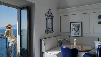 Une chambre avec un balcon donnant sur la mer, un canapé, une table avec des chaises et des tableaux encadrés au mur.