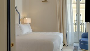 A hotel room with a bed, a chandelier, a lamp, a blue chair, and a view of a building through a window. - Hotel du Tasso (Sorrento)