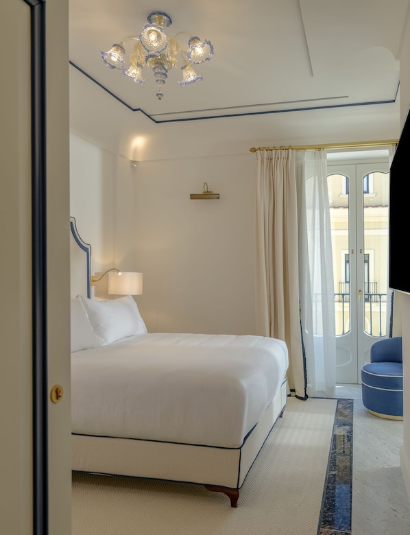 Habitación de hotel con cama, candelabro, lámpara, una silla azul y vista a un edificio a través de una ventana.