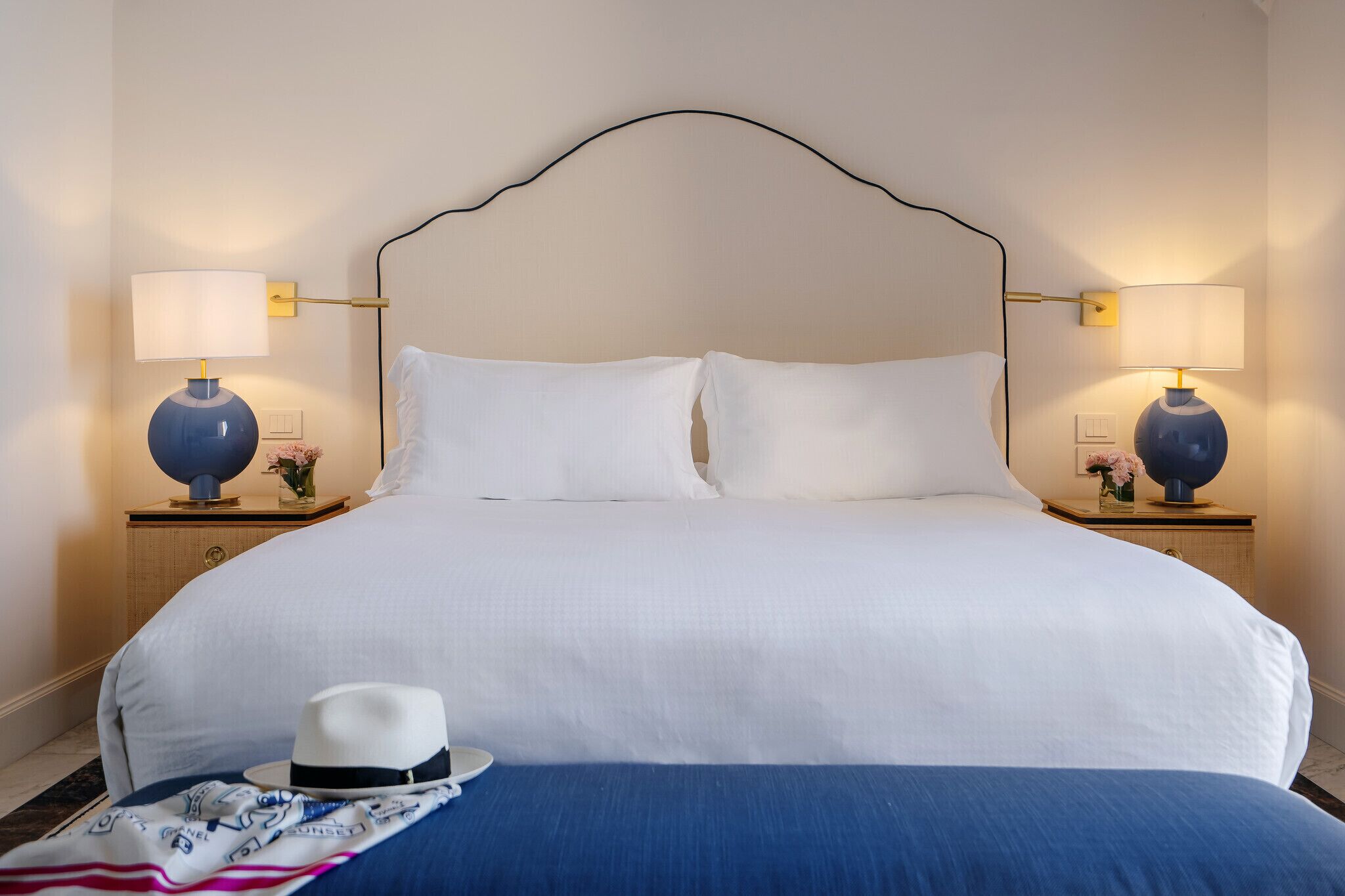 Una cama bien tendida con sábanas blancas, dos lámparas de noche, un sombrero decorativo y una manta azul sobre una mesita de madera.