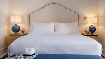 Una cama bien tendida con sábanas blancas, dos lámparas de noche, un sombrero decorativo y una manta azul sobre una mesita de madera.