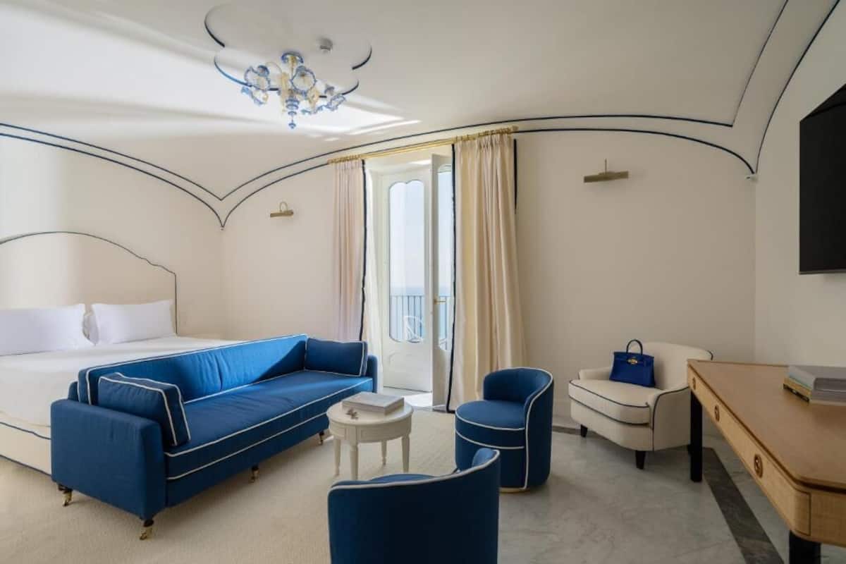 Habitación de hotel con una cama, un sofá azul, un sillón blanco, una mesita redonda pequeña y un escritorio de madera.