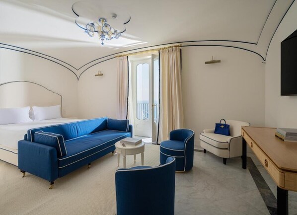 Habitación de hotel con una cama, un sofá azul, un sillón blanco, una mesita redonda pequeña y un escritorio de madera.