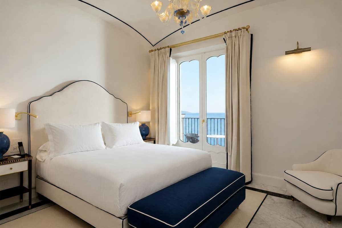 Un dormitorio con cama, mesitas de noche, un banco, una silla, una araña y vistas al mar.