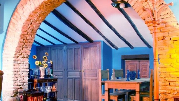 A room with a rustic brick archway, wooden ceiling beams, a wooden table with chairs, and a blue wall. - Tictágora House: House 2 (Plasenzuela)