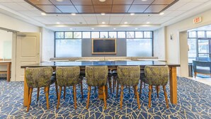 A conference room with a long table, six patterned chairs, a large screen, and a view of the outdoors through large windows.