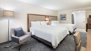 A hotel room with two beds, a desk, a chair, and a lamp.