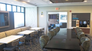 A modern hotel lobby with a long communal table, seating area, and a coffee station.