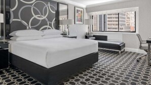 A modern hotel room with a large bed, a desk, a chair, and a view of the city through the window.