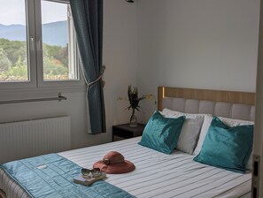 Ein Schlafzimmer mit Bett, Nachttisch, einem Fenster mit Vorhängen und Blick ins Grüne.