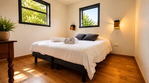 Ein Schlafzimmer mit einem Bett, zwei Fenstern, einem Nachttisch, einer Lampe und einer Zimmerpflanze.