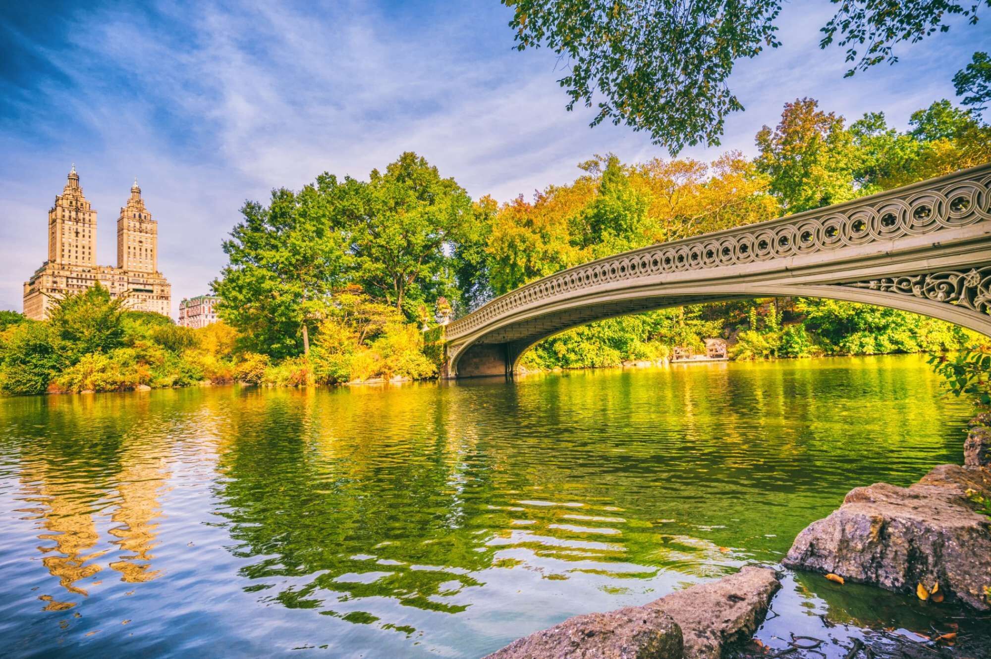 A stone bridge over a calm river with lush greenery and two prominent buildings in the background.