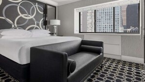 Una habitación de hotel moderna con una cama grande, un sofá negro, una mesita de noche con lámpara y un ventanal con vistas a la ciudad.