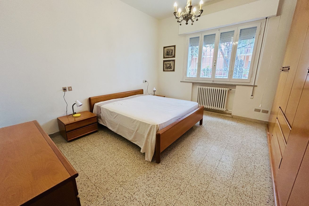 Ein Schlafzimmer mit Bett, Nachttisch, Holzkommode, Heizkörper und Fenster mit Blick auf Gebäude.