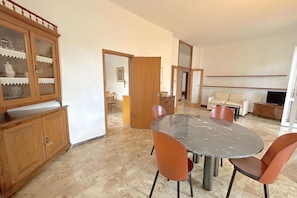 Een eethoek met een marmeren tafel en oranje stoelen, een houten kast met glazen deuren, en een bank op de achtergrond.
