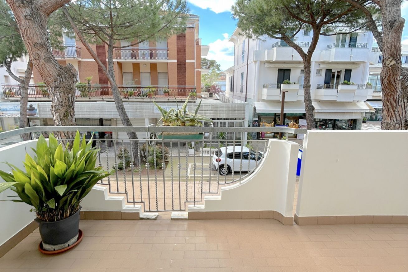 Un balcon orné d’un pot de fleurs, une voiture blanche garée au loin et des immeubles dotés de balcons et de fenêtres.