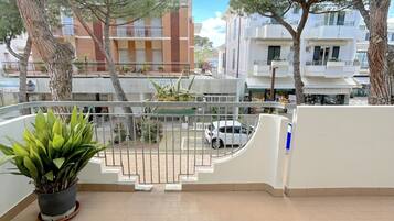 Un balcon orné d’un pot de fleurs, une voiture blanche garée au loin et des immeubles dotés de balcons et de fenêtres.
