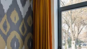 Una habitación con pared decorada, una ventana con cortinas amarillas, una mesa con una taza de té, y vistas a árboles y una calle.