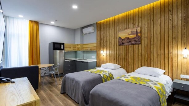 Habitación de hotel con dos camas, un escritorio, una silla, un refrigerador y una pared de madera con una imagen de la ciudad.