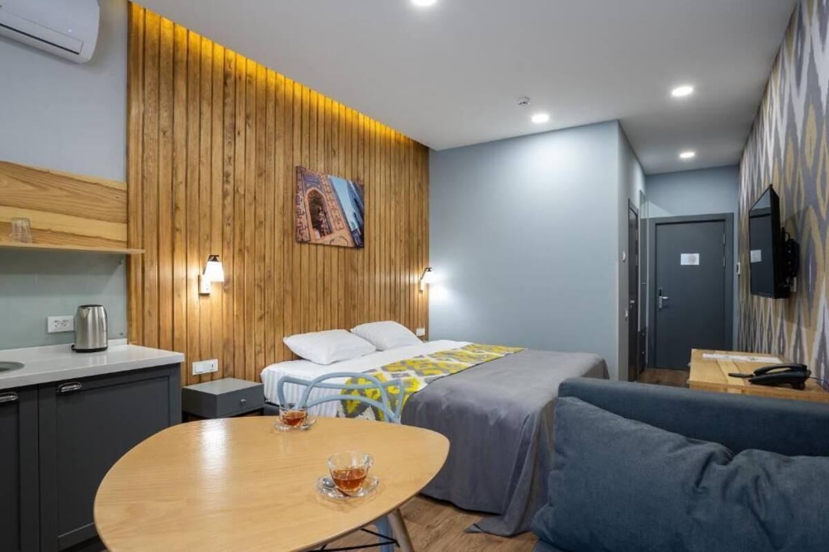 A modern hotel room with a wooden accent wall, a bed with a blue and yellow bedspread, a small table with a cup, a sofa, a TV, and a kitchenette.