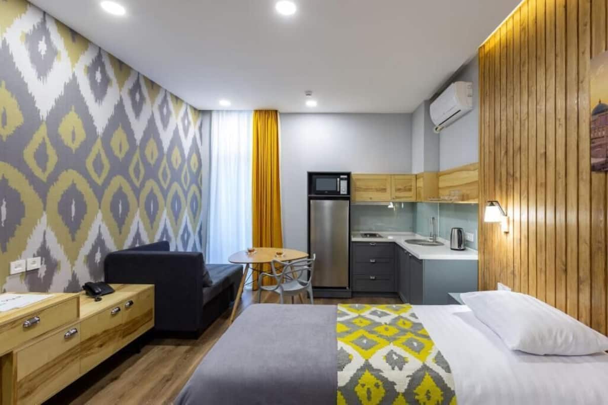A compact hotel room with a bed, a small desk, a kitchenette, and a seating area.