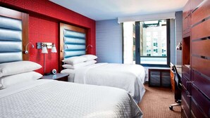 Hotellihuone, jossa on kaksi sänkyä, punainen, geometrisesti kuvioitu seinä, suuri ikkuna, josta on näkymä kaupunkiin, ja puinen vaatekaappi.