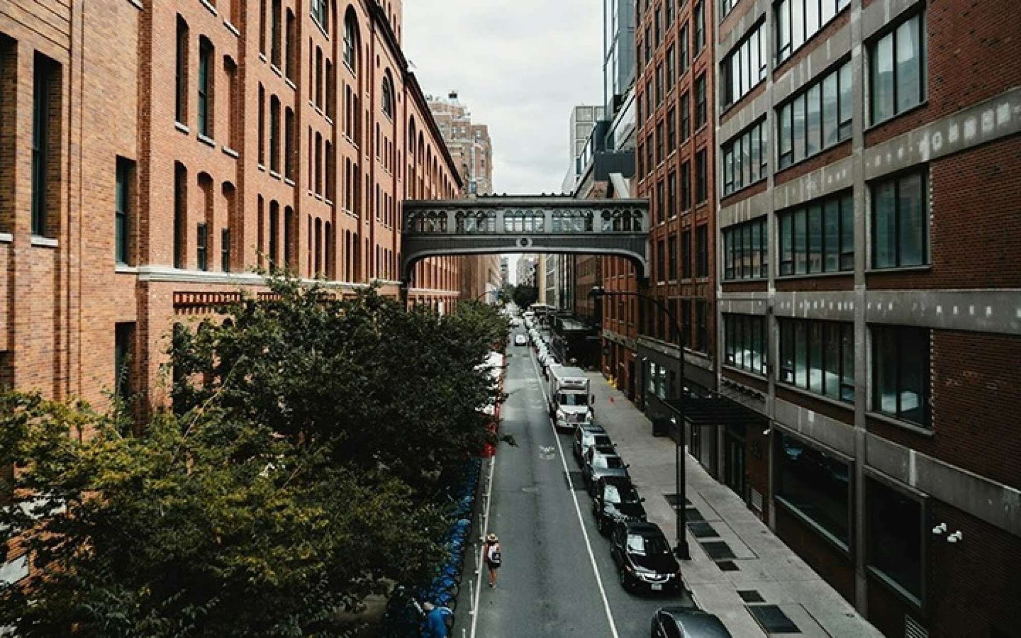 Vista de una calle con un puente peatonal, edificios, coches aparcados y árboles.