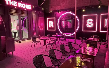 An outdoor seating area with tables and chairs, neon signage, and a glass door entrance.