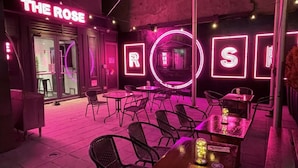 An outdoor seating area with tables and chairs, neon signage, and a glass door entrance.
