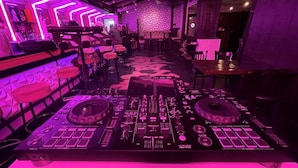 A DJ booth with a control panel and two turntables, surrounded by bar seating and a patterned wall.