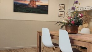 Un comedor con una mesa de madera y dos sillas blancas, una planta en maceta con flores y un cuadro de una iglesia en la pared.