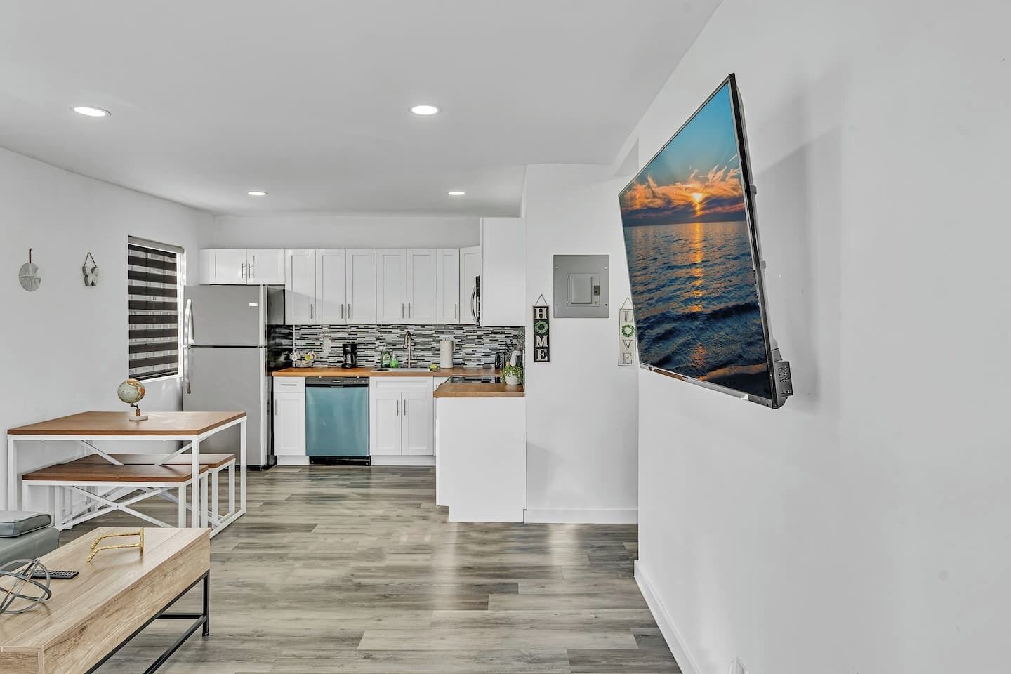 A modern kitchen with white cabinets, a wooden countertop, and a dishwasher. There is a dining area with a wooden table and chairs. A large TV is mounted on the wall displaying a sunset over the ocean.