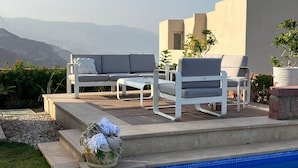 Zona junto a la piscina con sillas de descanso, una mesita y vistas a las montañas.