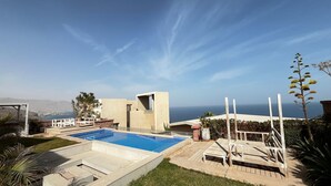 Un área de piscina moderna al aire libre con vista despejada al océano, un pequeño edificio y una terraza de madera con sillas.