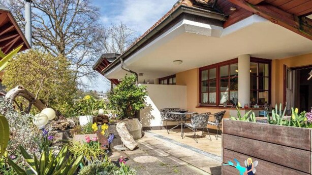 Une terrasse avec une table et des chaises, entourée d’un jardin où poussent différentes plantes et fleurs.