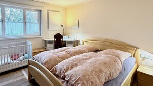 Une chambre à coucher avec un lit double, un bureau, une chaise, un berceau et une fenêtre donnant sur des arbres.