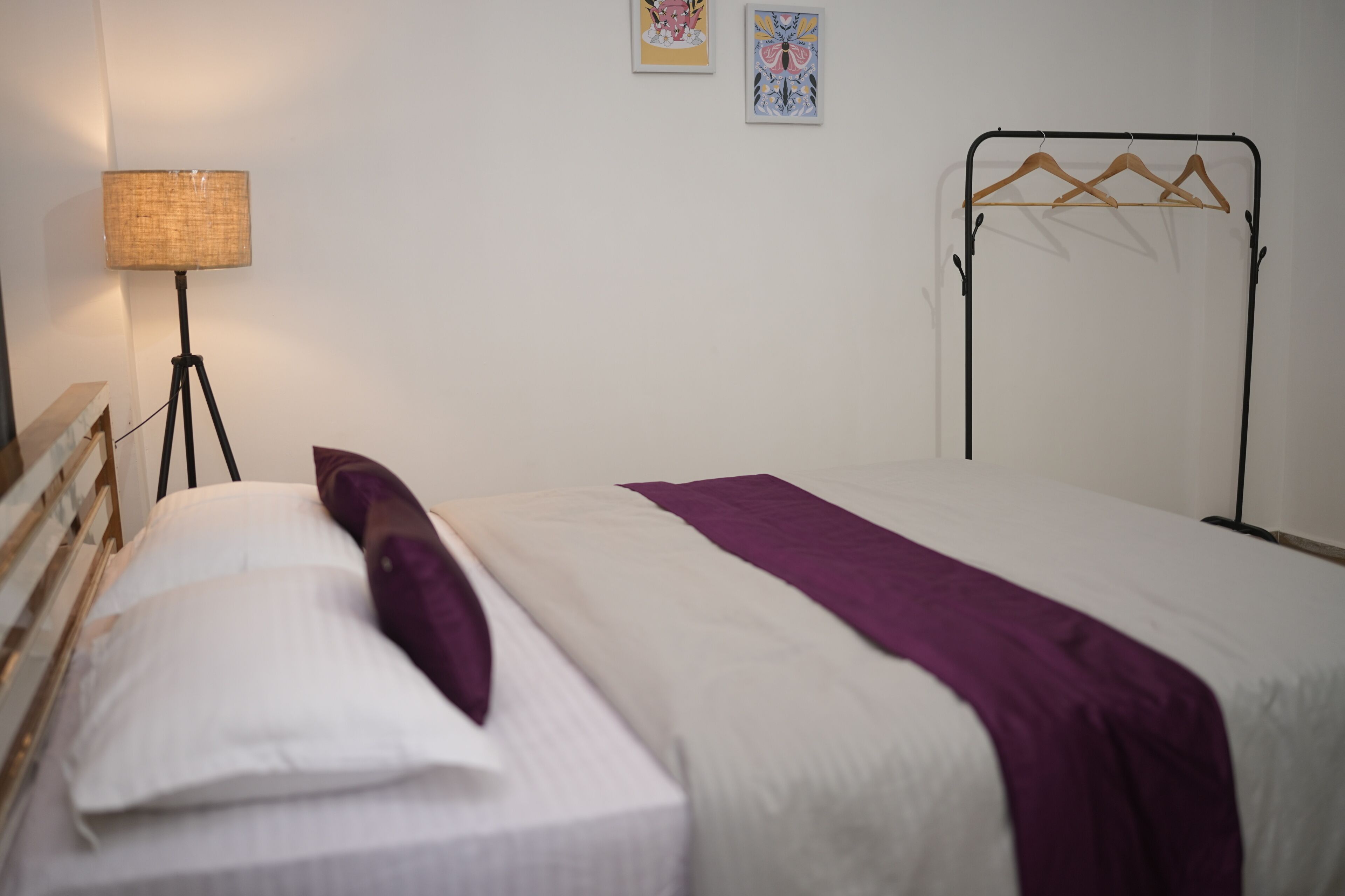 A neatly made bed with a white and purple bedspread, a single white pillow, and a floor lamp with a woven shade.