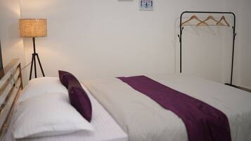 A neatly made bed with a white and purple bedspread, a single white pillow, and a floor lamp with a woven shade.