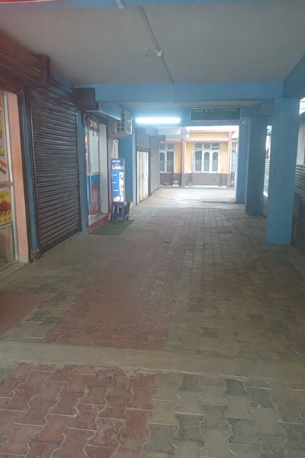 A corridor with closed shops on both sides, a tiled floor, and a sign for 'ONE EYE JYOTISH' in the distance. - Shivalik stays (East Jorhat)