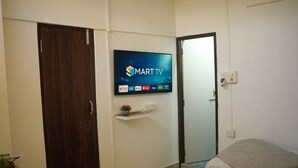 Une chambre à coucher avec un téléviseur fixé au mur, affichant l’interface d’un service de diffusion en continu, une porte en bois, une table de chevet avec une plante et un lit recouvert d’une couette blanche.