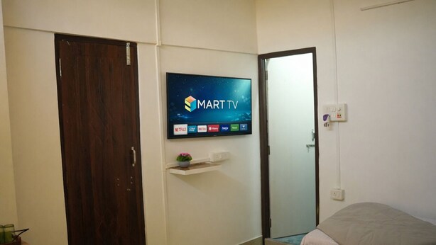A bedroom with a TV mounted on the wall displaying a streaming service interface, a wooden door, a bedside table with a plant, and a bed with a white comforter. - Shivalik stays  (East Jorhat)