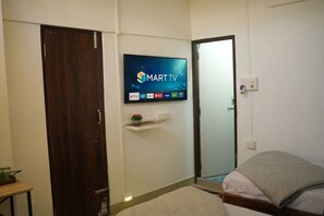 A bedroom with a TV mounted on the wall displaying a streaming service interface, a wooden door, a bedside table with a plant, and a bed with a white comforter. - Shivalik stays (East Jorhat)