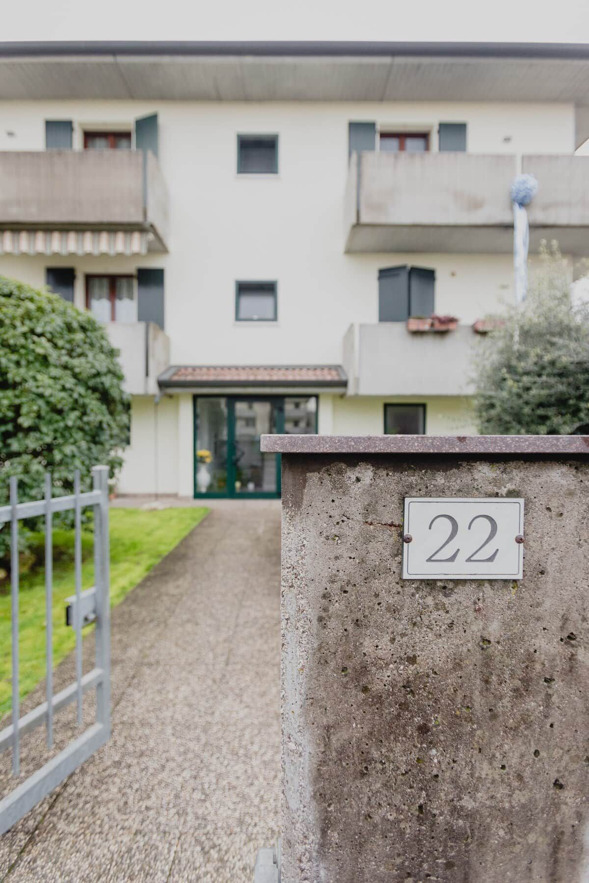 A building entrance with a concrete wall displaying the number 22, a gravel path leading to the entrance, and a metal gate.